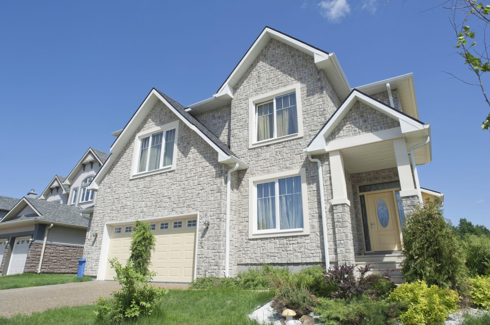 Modern two-story suburban house with stone exterior, large windows, attached garage, and landscaped front yard under a clear blue sky.