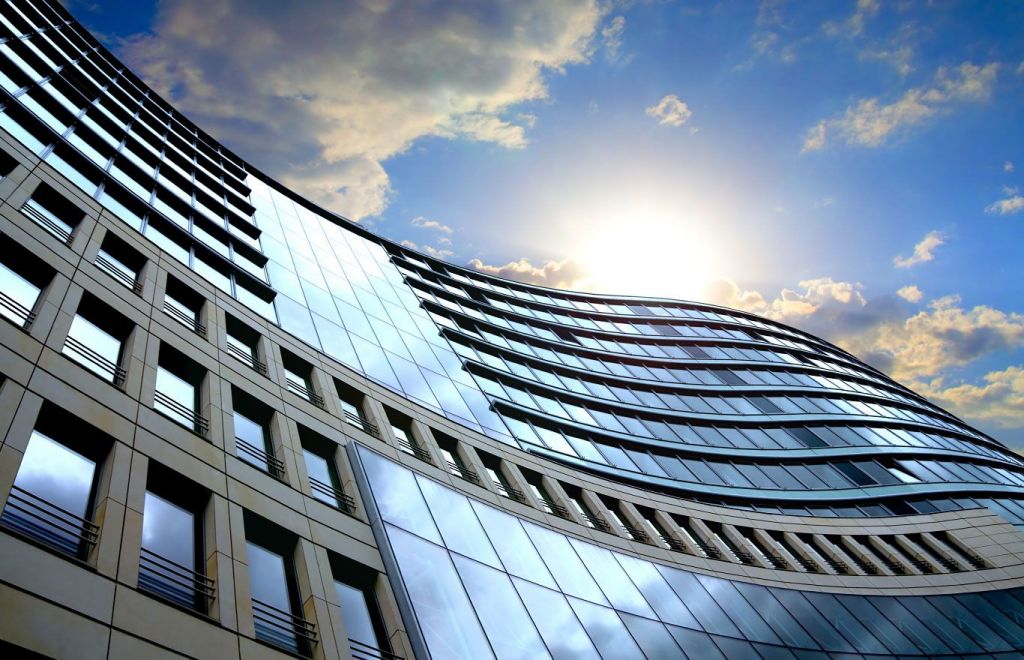 Low angle view of a modern commercial building with a curved glass facade and sunburst against a blue sky.